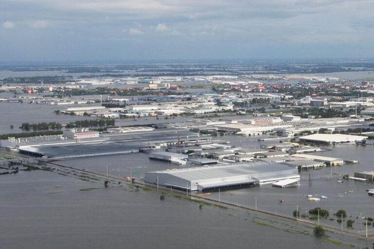 Veduta aerea di un'area industriale colpita dall'alluvione, con edifici e strade parzialmente sommersi dall'acqua; sullo sfondo sono visibili il paesaggio urbano lontano e il cielo nuvoloso.