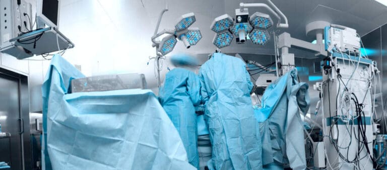 Chirurghi in camice e berretto blu eseguono un'operazione in una sala operatoria moderna e luminosa con attrezzature mediche avanzate, evidenziando l'importanza dell'assicurazione di viaggio e del rimborso spese mediche per gli espatriati durante il ricovero d'emergenza.