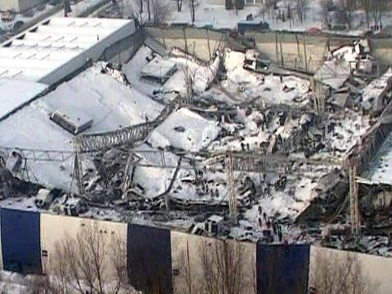 Un grande edificio con il tetto coperto di neve è parzialmente crollato, lasciando metallo contorto e detriti sparsi sul posto. Intorno alla struttura sono visibili diversi veicoli e in primo piano ci sono degli alberi.