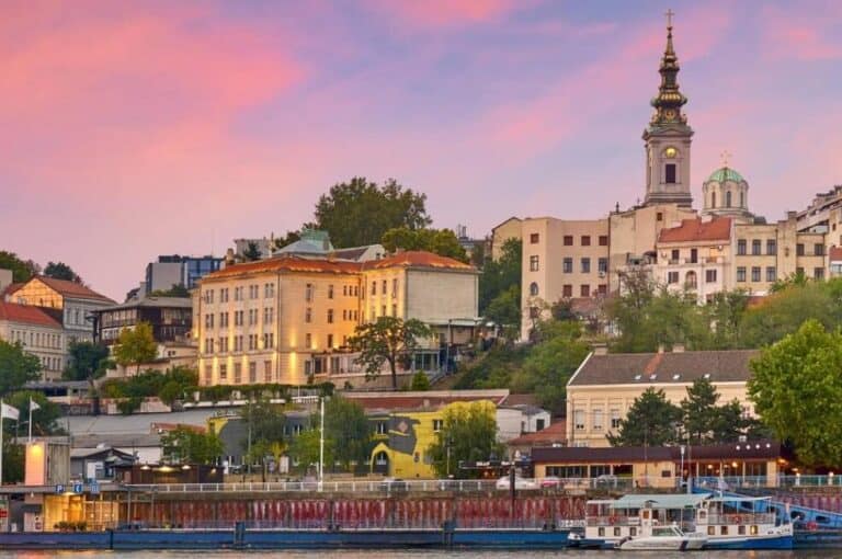 Una veduta fluviale di una città serba con edifici storici, una chiesa importante con un'alta guglia, alberi e una barca attraccata al tramonto sotto un cielo rosa e viola.