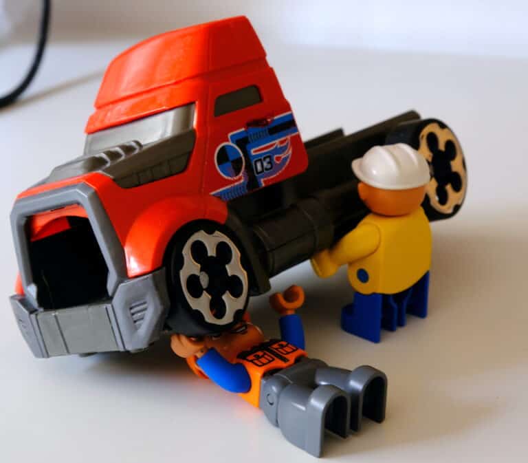 Due figure giocattolo lavorano su un camion giocattolo; una con un cappello rigido sta accanto al camion, mentre l'altra è sdraiata sotto come se stesse riparando un vizietto difettoso. Il camion rosso e grigio è appoggiato su una superficie bianca.