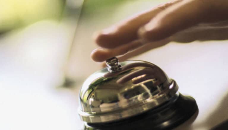 Un primo piano di una mano che preme su un campanello di servizio lucido, tipico delle reception degli hotel, con uno sfondo sfocato, a simboleggiare la pronta attenzione e la riduzione dei rischi per gli ospiti.