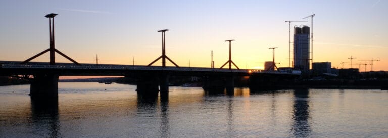 Un ponte attraversa un fiume tranquillo al tramonto, con edifici moderni e gru edili sullo sfondo. Il cielo si illumina di luce calda e i riflessi scintillano sull'acqua: una scena serena spesso ammirata da un amministratore dopo il trasferimento all'estero.