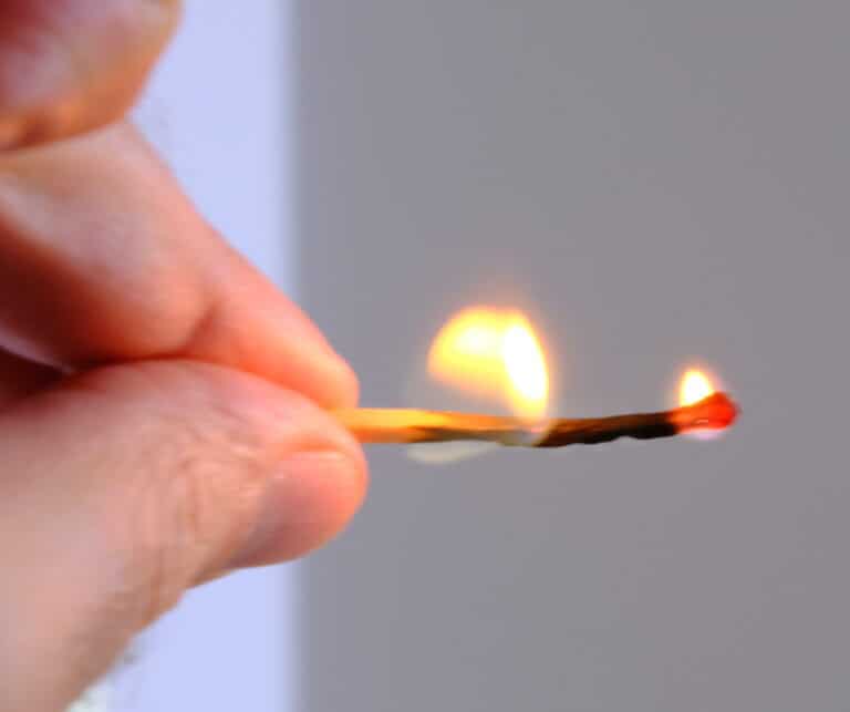 Primo piano di una mano che tiene un fiammifero acceso con una piccola fiamma sulla punta, simbolo di rischio patrimonio personale per amministratore di aziende estere, su uno sfondo neutro sfocato.