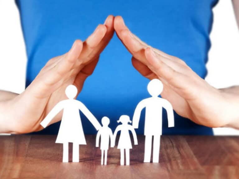 Le mani di una persona formano un tetto protettivo su ritagli di carta di una famiglia con due adulti e due bambini, a simboleggiare la protezione, la cura a lungo termine e la salvaguardia dei risparmi.