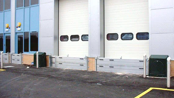 Baia di carico con due porte a rullo industriali bianche chiuse, barriere protettive, cassette di servizio verdi e linee di parcheggio gialle su una superficie pavimentata all'esterno di un edificio commerciale progettato per resistere ai rischi di alluvione.