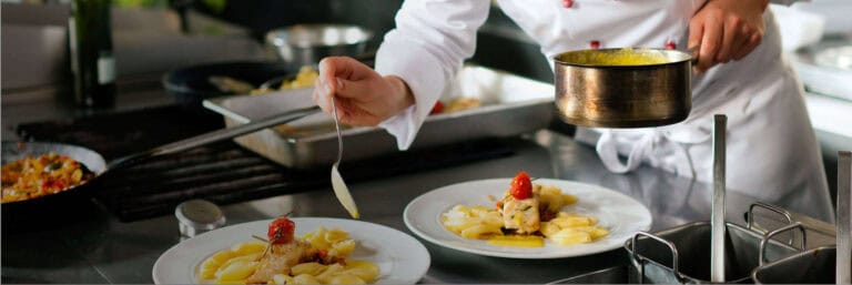 Uno chef in uniforme bianca versa la salsa su piatti gourmet con carne, patate e una guarnizione di pomodoro in una cucina professionale, facendo attenzione a evitare infezioni o avvelenamento alimentare.