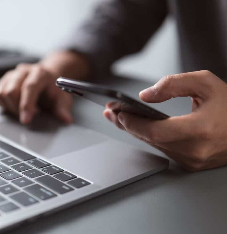 Una persona che utilizza uno smartphone con una mano mentre con l'altra digita sulla tastiera di un computer portatile, seduta a una scrivania con uno sfondo sfocato.