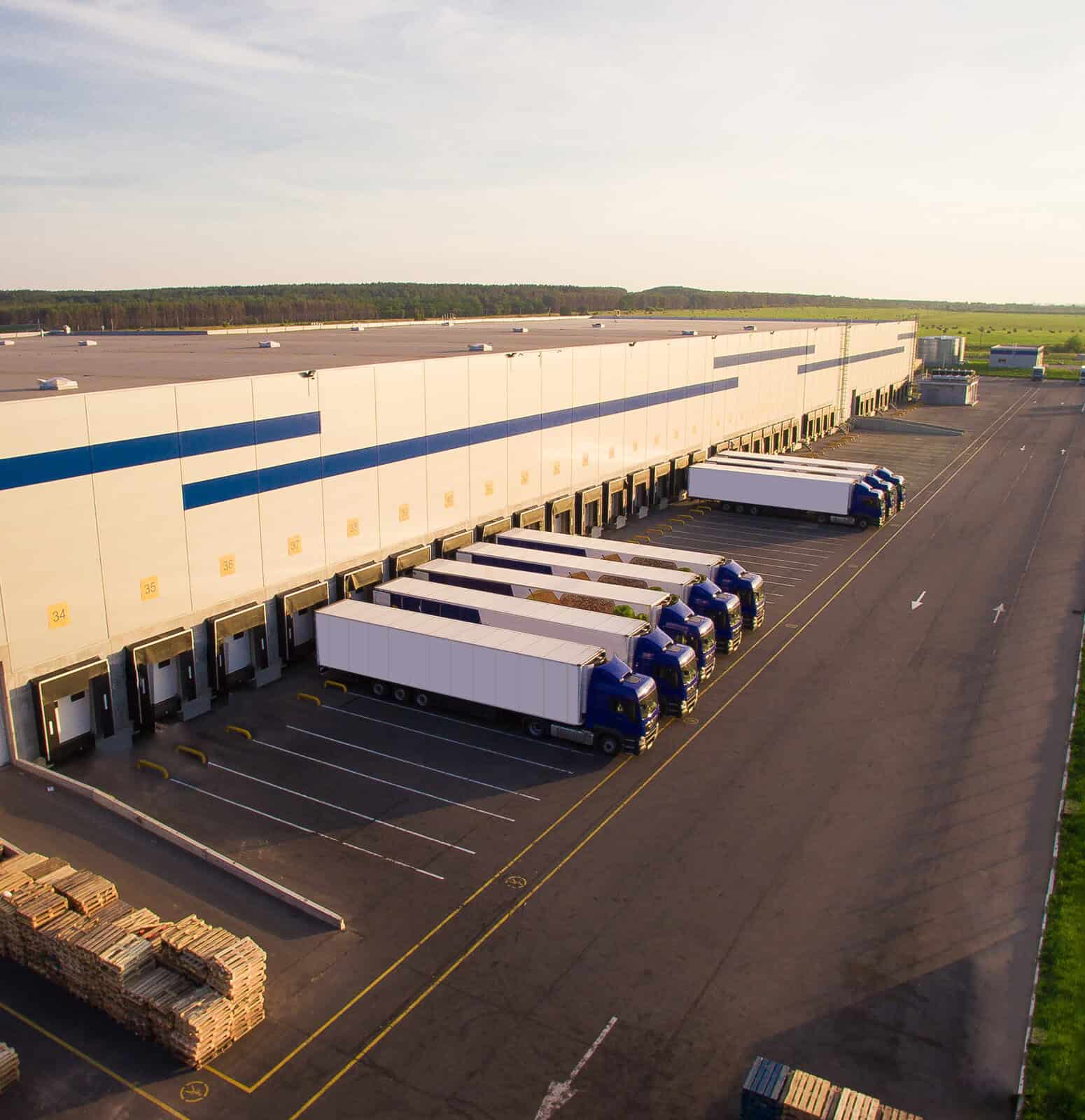 Aerial view of a large warehouse with several loading docks where multiple trucks are parked for loading or unloading, surrounded by paved areas and stacks of pallets.