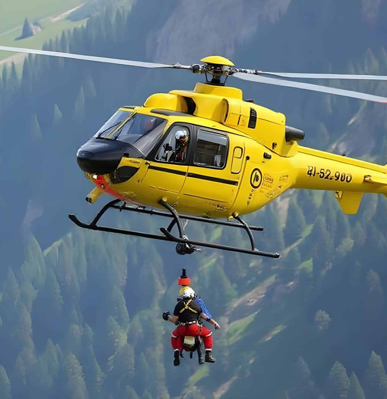 A yellow rescue helicopter hovers above a wooded mountain slope while lifting a person with a “harness during an” aerial rescue operation.