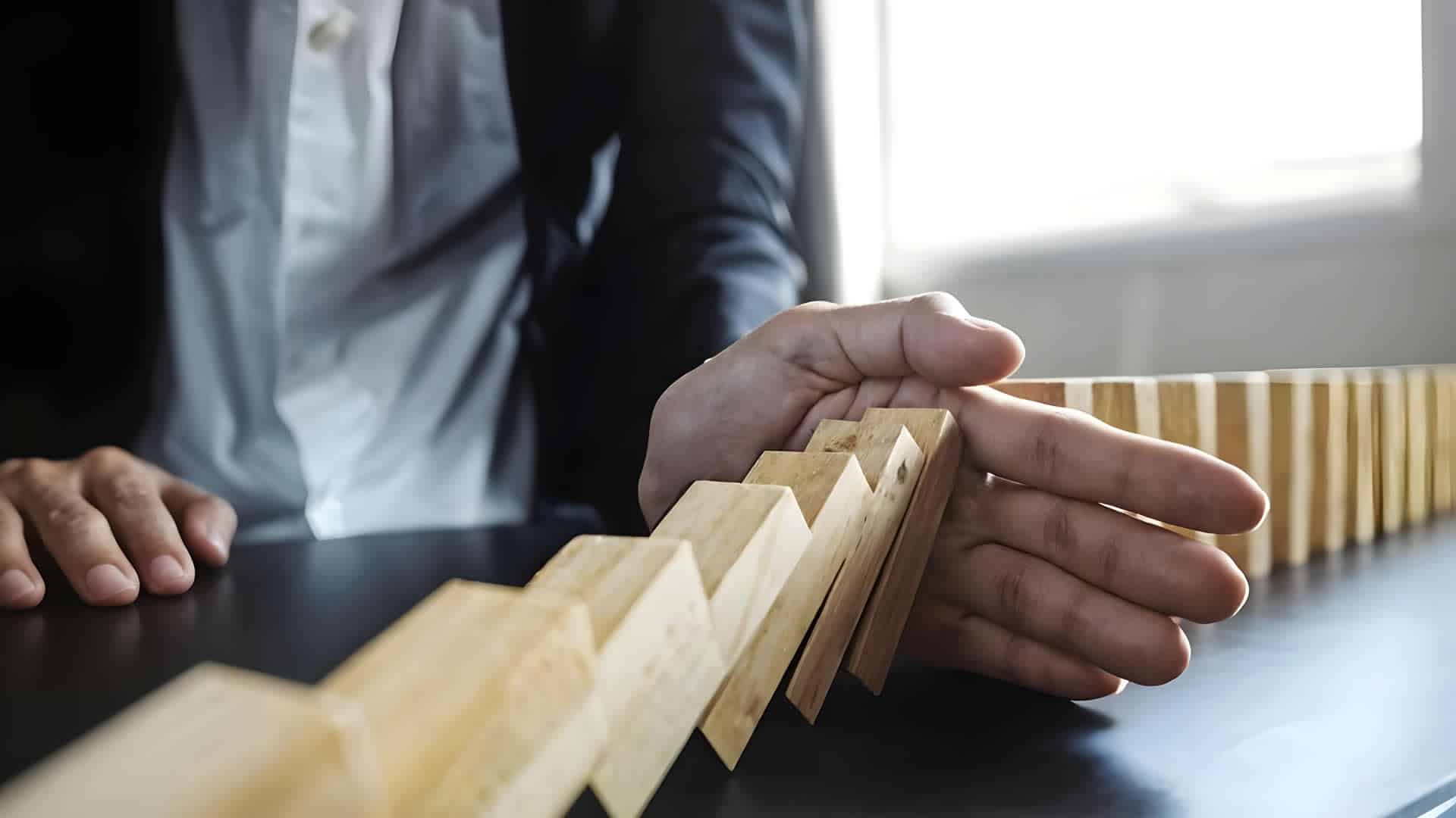 A person in a suit uses their hand to stop a line of falling wooden dominoes on a dark table, preventing the rest from toppling over.