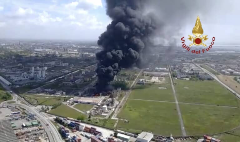 Aerial view of a large industrial fire with thick black smoke rising into the sky; surrounding area includes fields, buildings, and shipping containers. The Vigili del Fuoco (Italian fire brigade) emblem is visible.