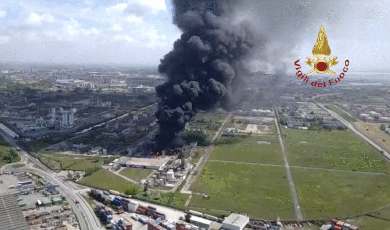 Veduta aerea di un grande incendio industriale con un denso fumo nero che sale verso il cielo; il logo dei Vigili del Fuoco è visibile nell'angolo in alto a destra.